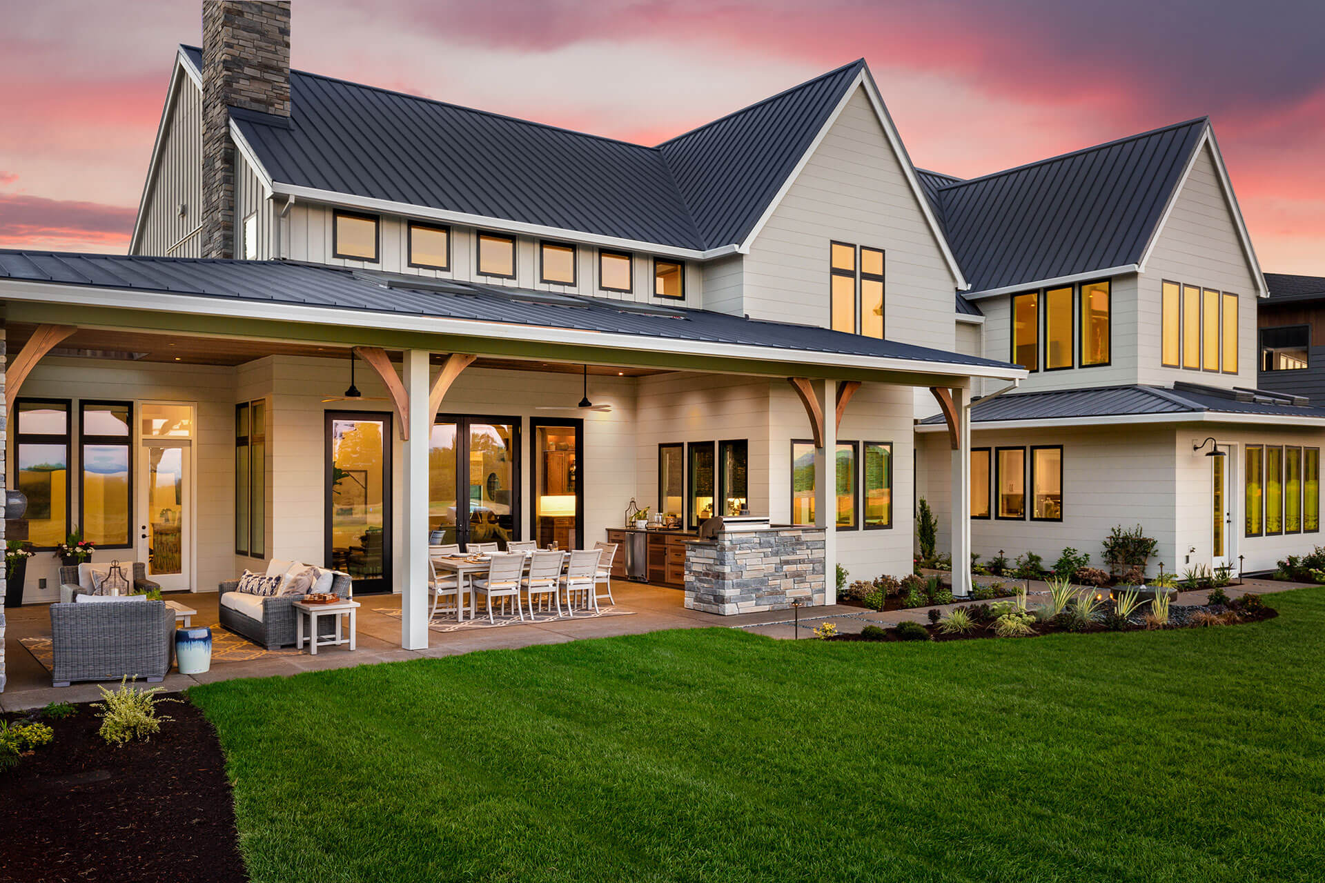 New luxury home exterior at sunset. Features large covered patio with barbecue, outdoor kitchen, and furniture